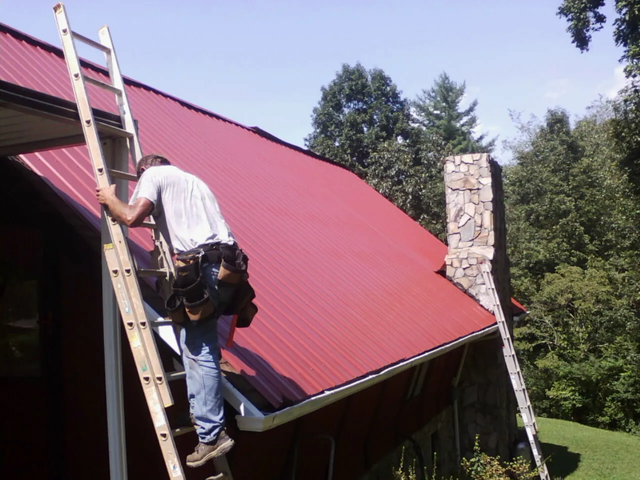 Roofer on a ladder beside a red metal roof and stone chimney — Soffit & Fascia Repair by Roofing Brothers in Bethany